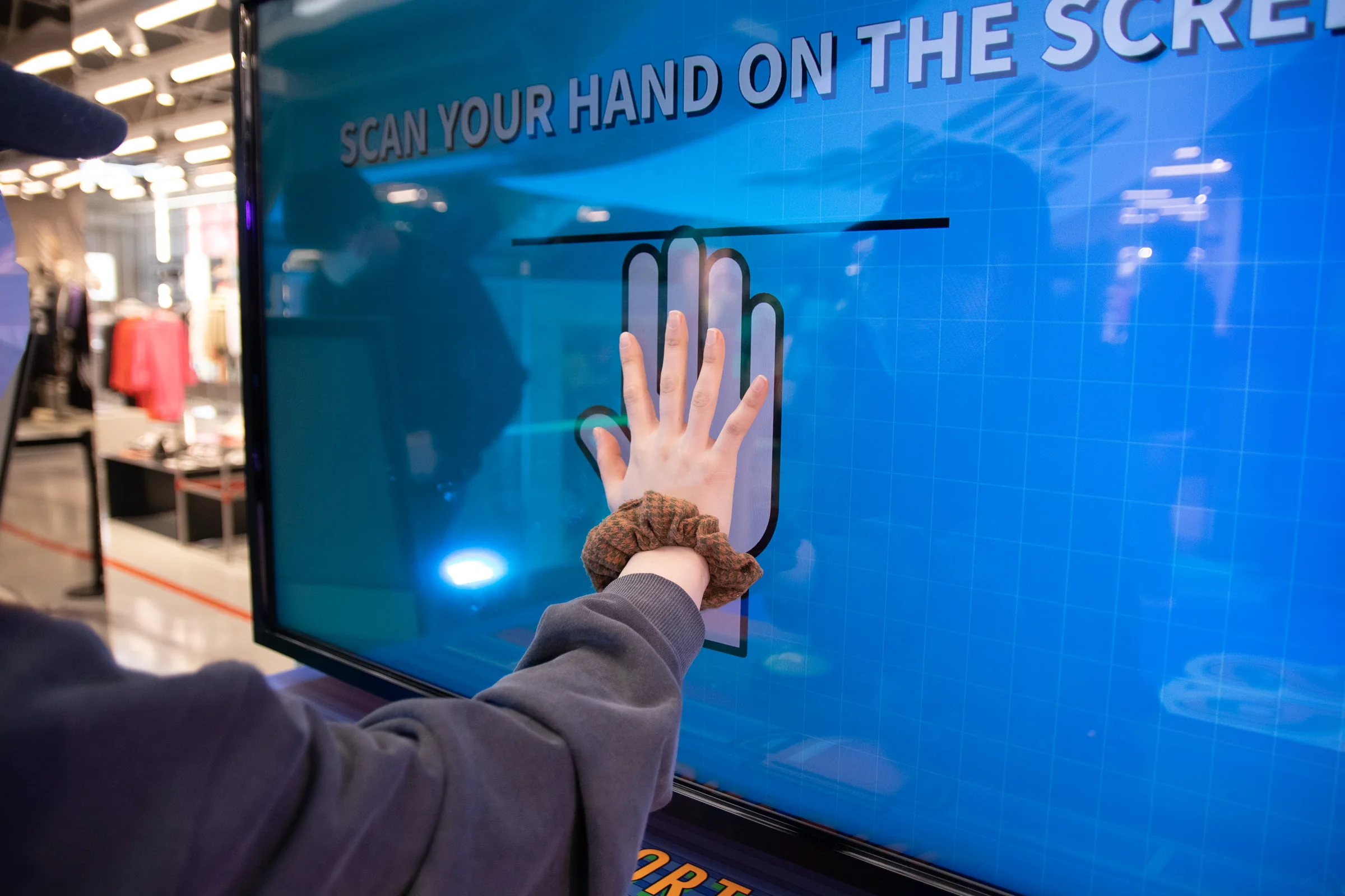 A participant places a palm on the screen displaying SCAN YOUR HAND ON THE SCREEN.