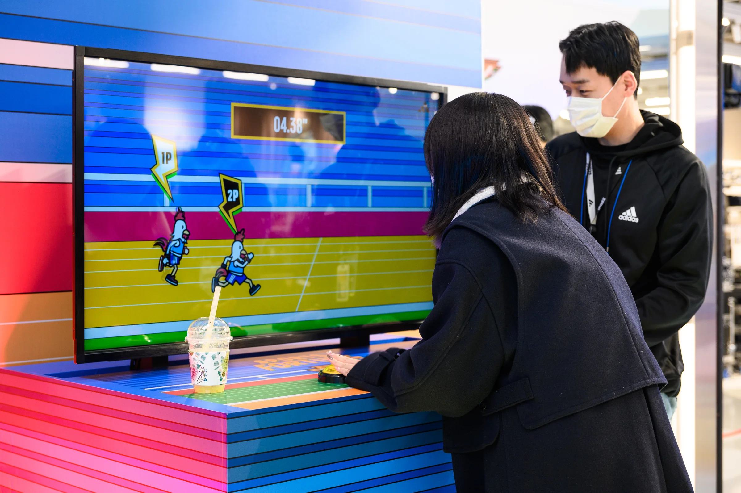 A participant presses buttons during a two-player race showing 04.38 seconds, with staff watching nearby.