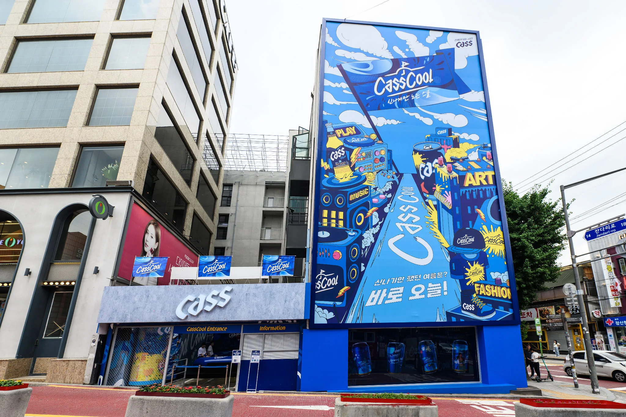 모퉁이에서 본 메인 팝업. 낮은 건물의 Cass 간판 입구 옆으로 카스쿨 벽화가 그려진 높은 건물이 서 있다.
