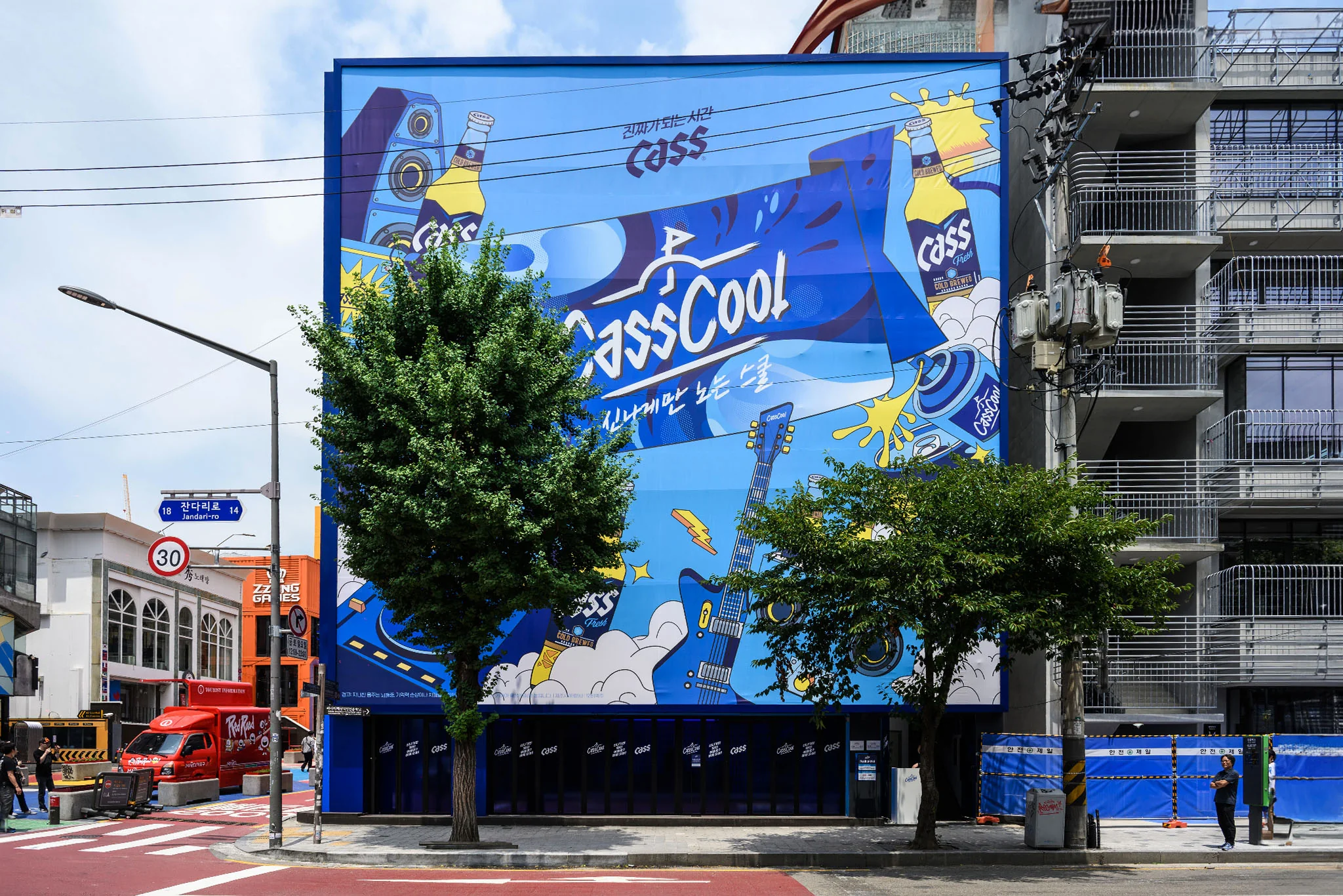 건물 한 면을 덮은 카스쿨 벽화. 파란 바탕에 CassCool 글자와 기타, 카스 캔이 그려져 있고 앞에 가로수가 서 있다.