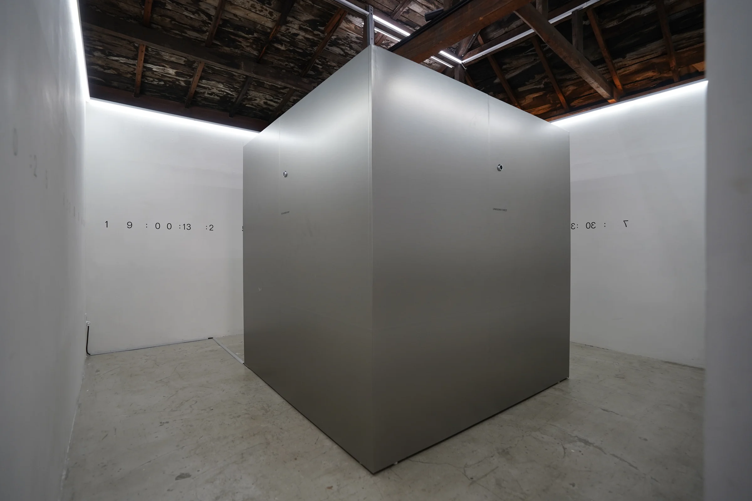 A metal cube stands in a white room beneath an old timber ceiling.