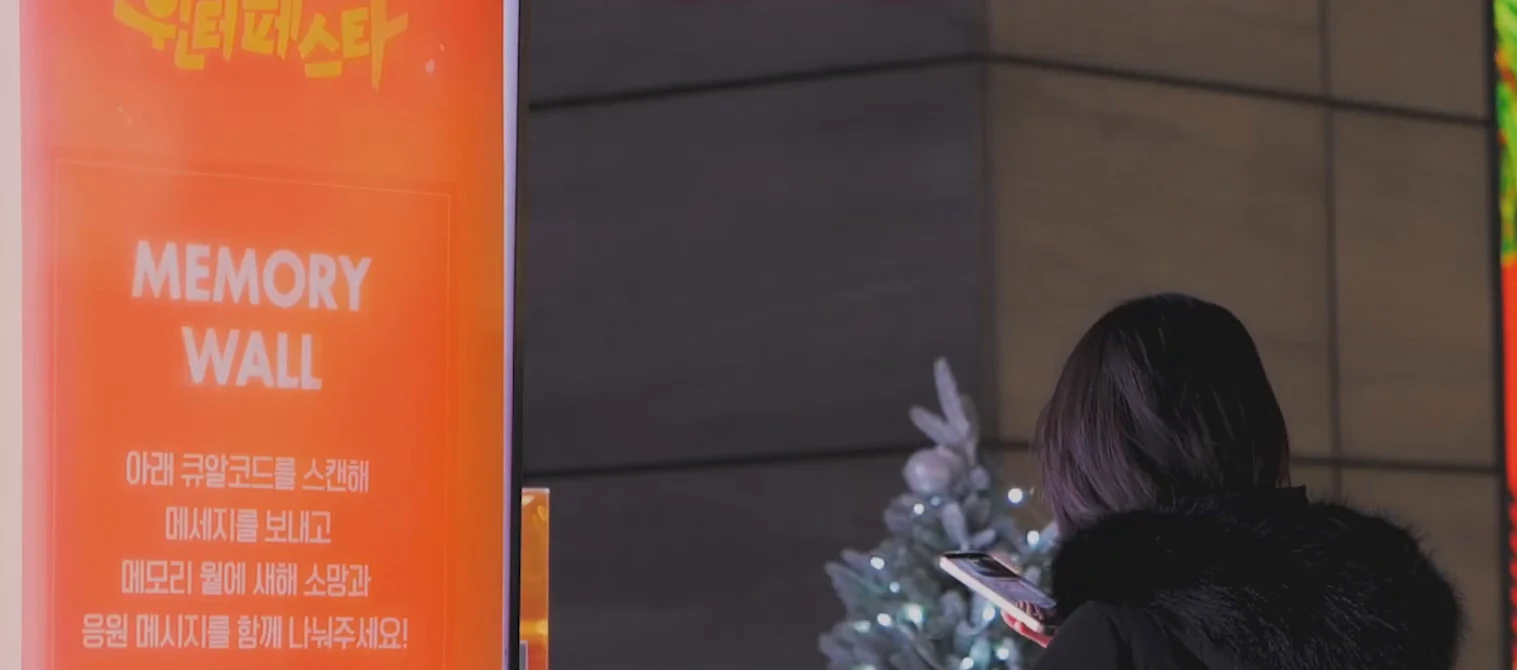 MEMORY WALL 안내 배너 옆에서 휴대폰으로 QR을 비추는 관객