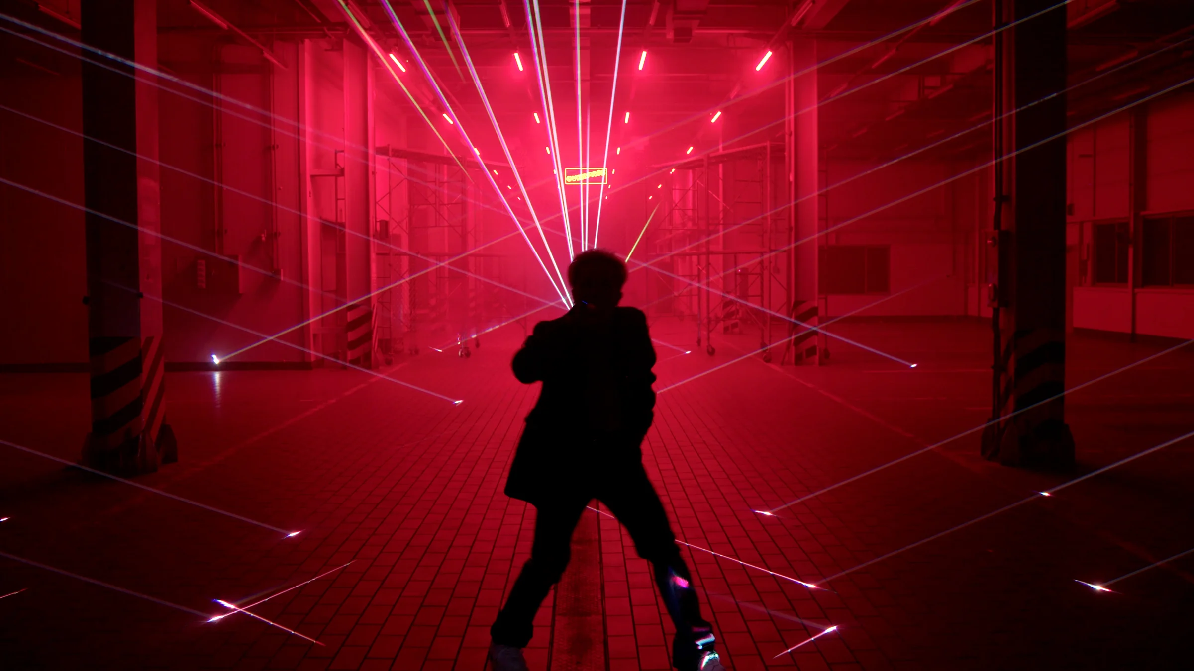 A silhouette at the center of the warehouse with red beams radiating outward